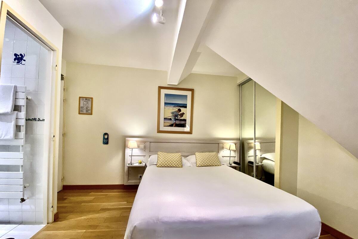 Ein Hotelzimmer mit einem großen Bett, Holzboden, einem Gemälde an der Wand und einem Badezimmer mit weißer Tür.