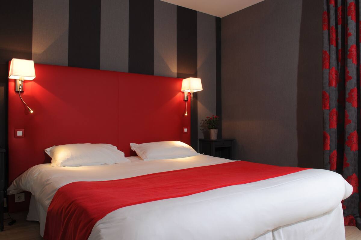 Ein Hotelzimmer mit einem großen Bett, einem roten Kopfteil, zwei Nachttischlampen und einem dunklen Nachttisch mit einer Topfpflanze.