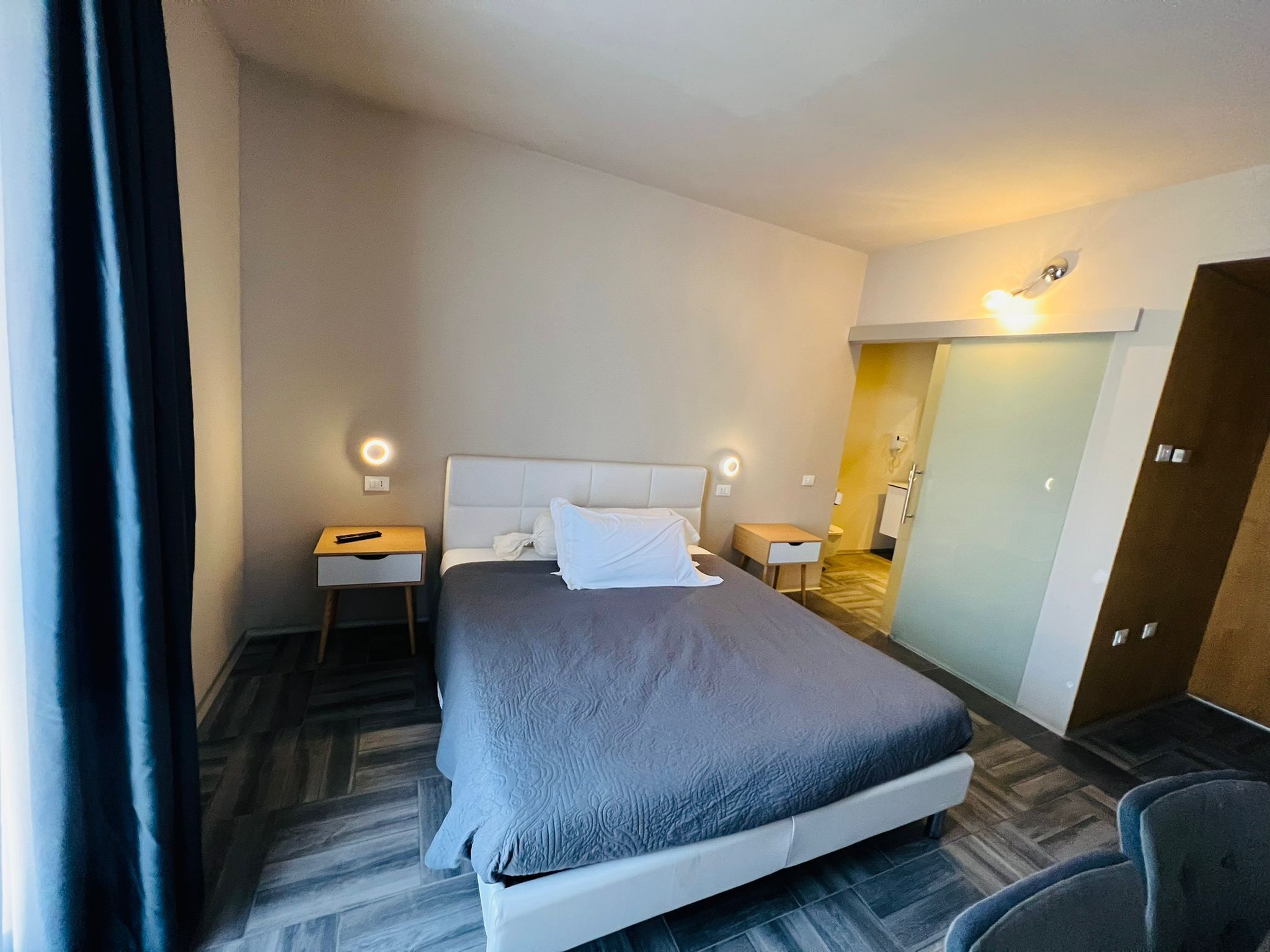 A hotel room with a large bed, two bedside tables, a nightstand, a chair, and a wardrobe.