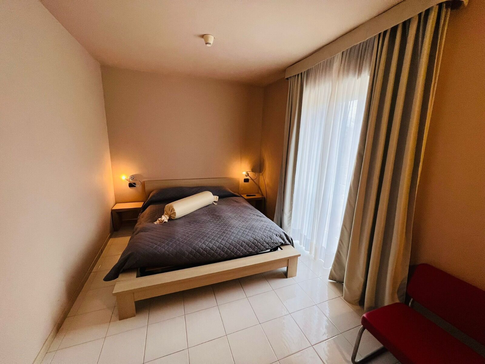 Een slaapkamer met een bed, nachtlampjes, een rode stoel en een raam met gordijnen.