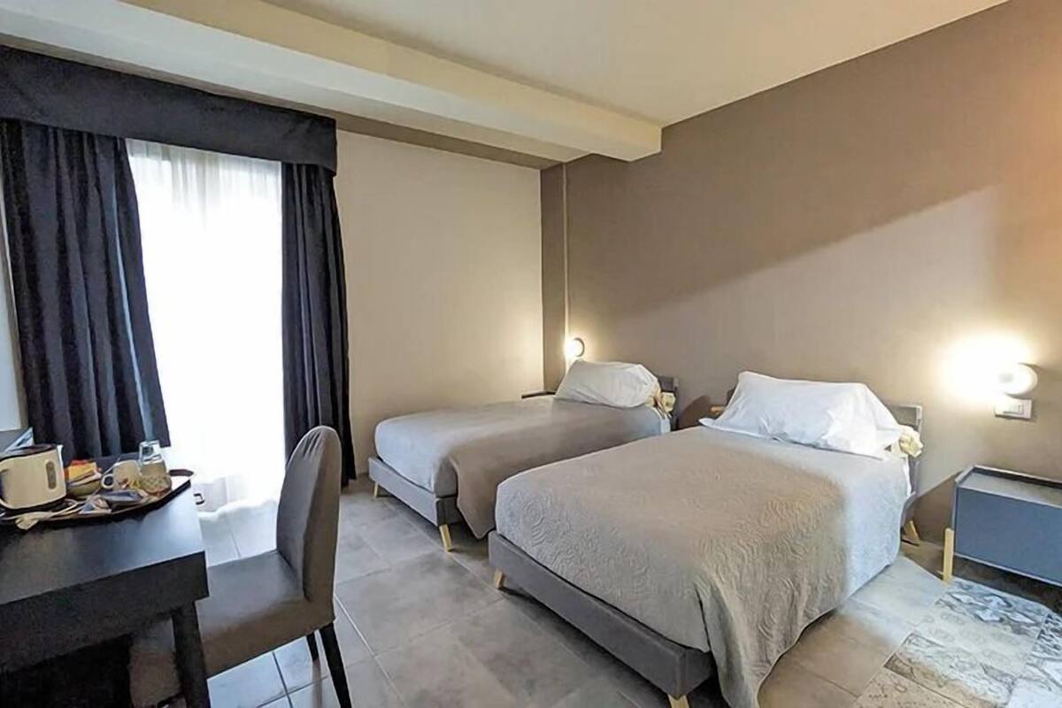 A hotel room with two single beds, a desk with a chair, a small table with a tray of items, and a window with curtains.