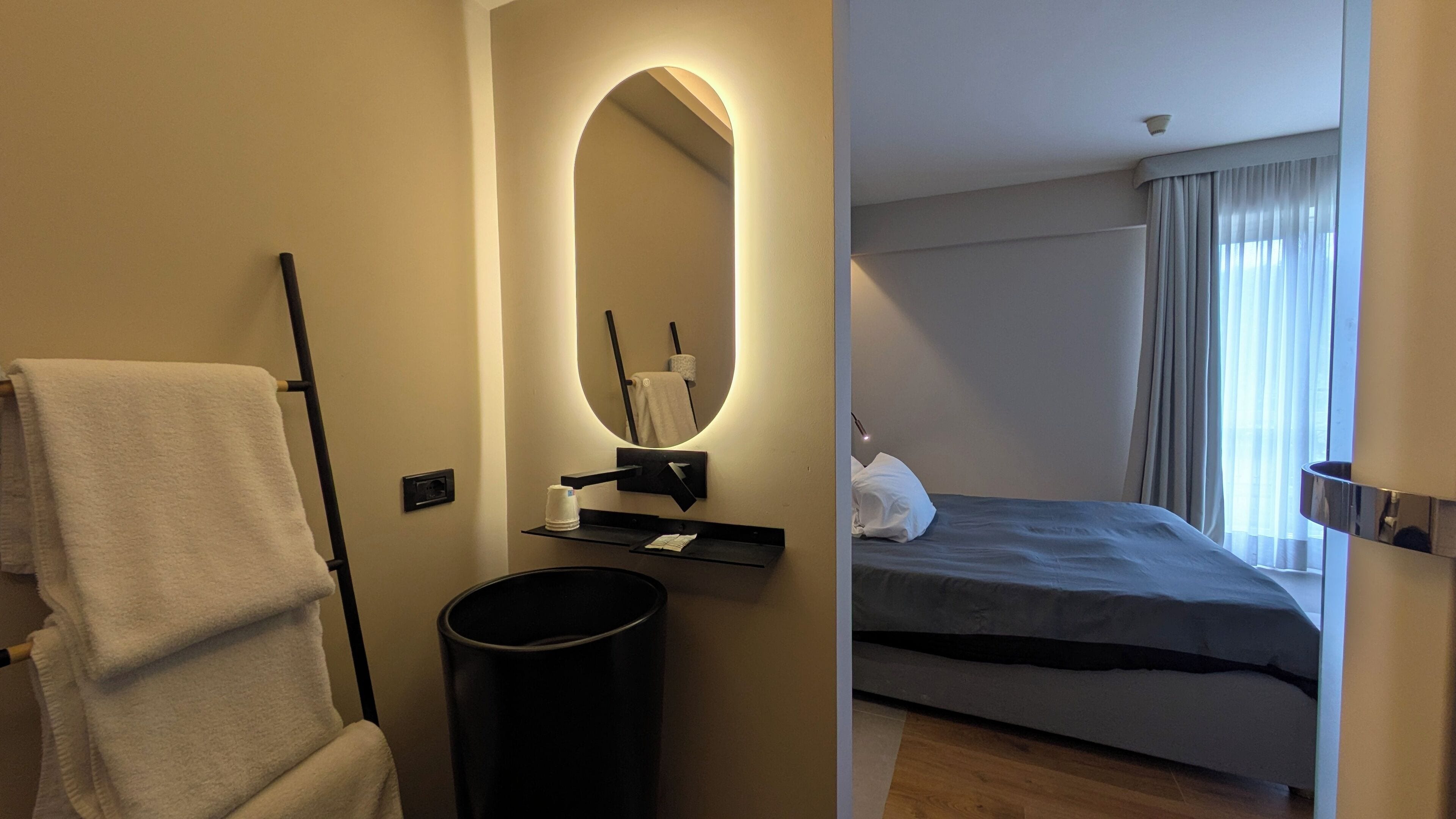 A hotel room with a large bed, a bedside table, a mirror, a towel rack, and a trash bin.