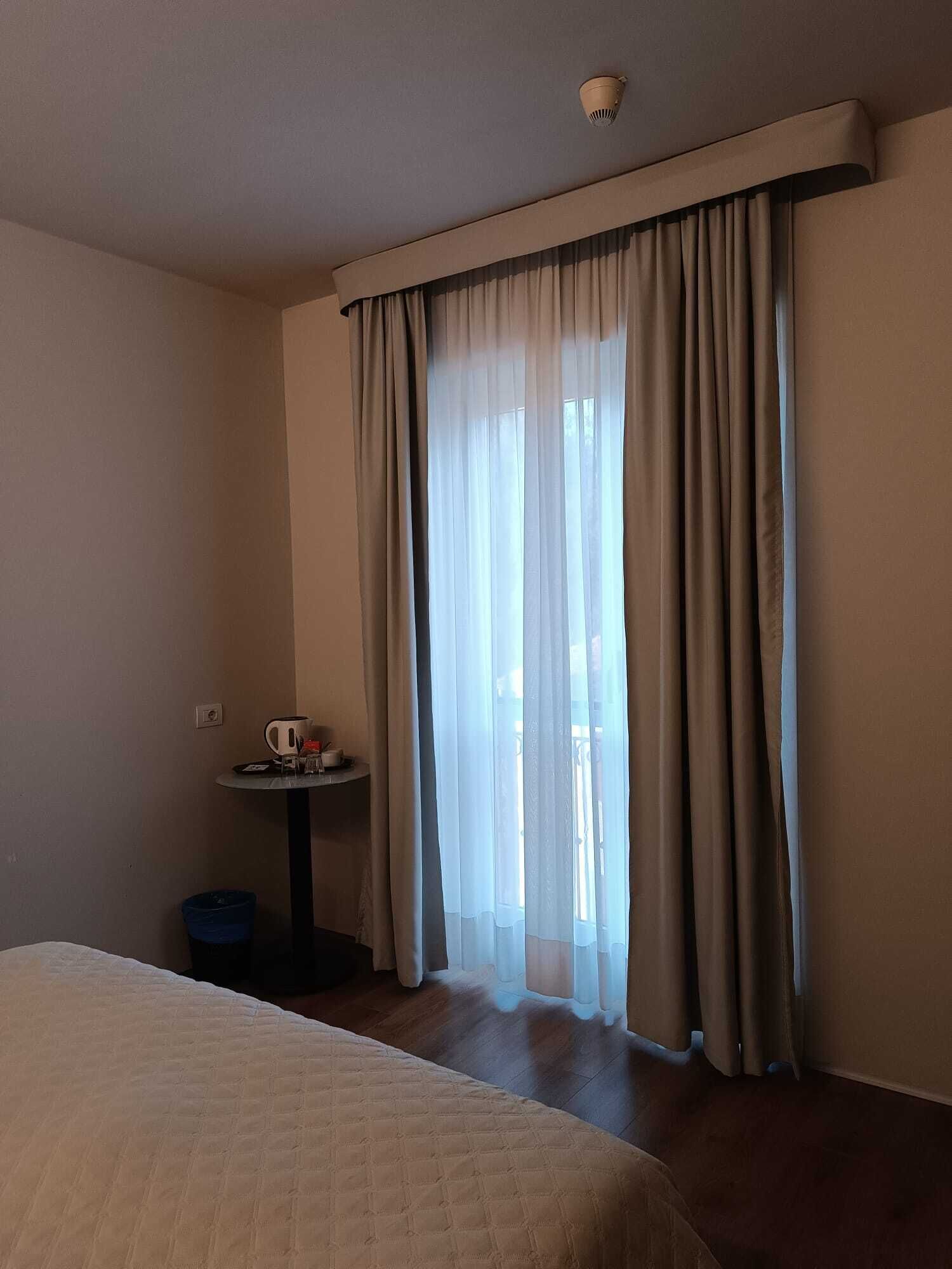 A hotel room with a bed, a small table with a kettle and cups, and a window with curtains.
