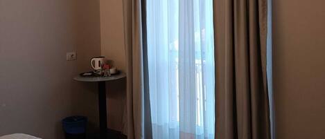 A hotel room with a bed, a small table with a kettle and cups, and a window with curtains.