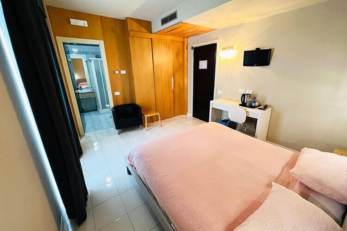 A hotel room with a bed, a desk, a chair, a TV, and a bathroom visible through an open door.