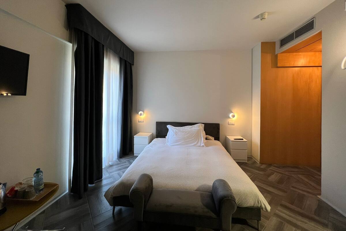 A hotel room with a large bed, two bedside tables, a flat-screen TV, a window with curtains, and a wooden door.