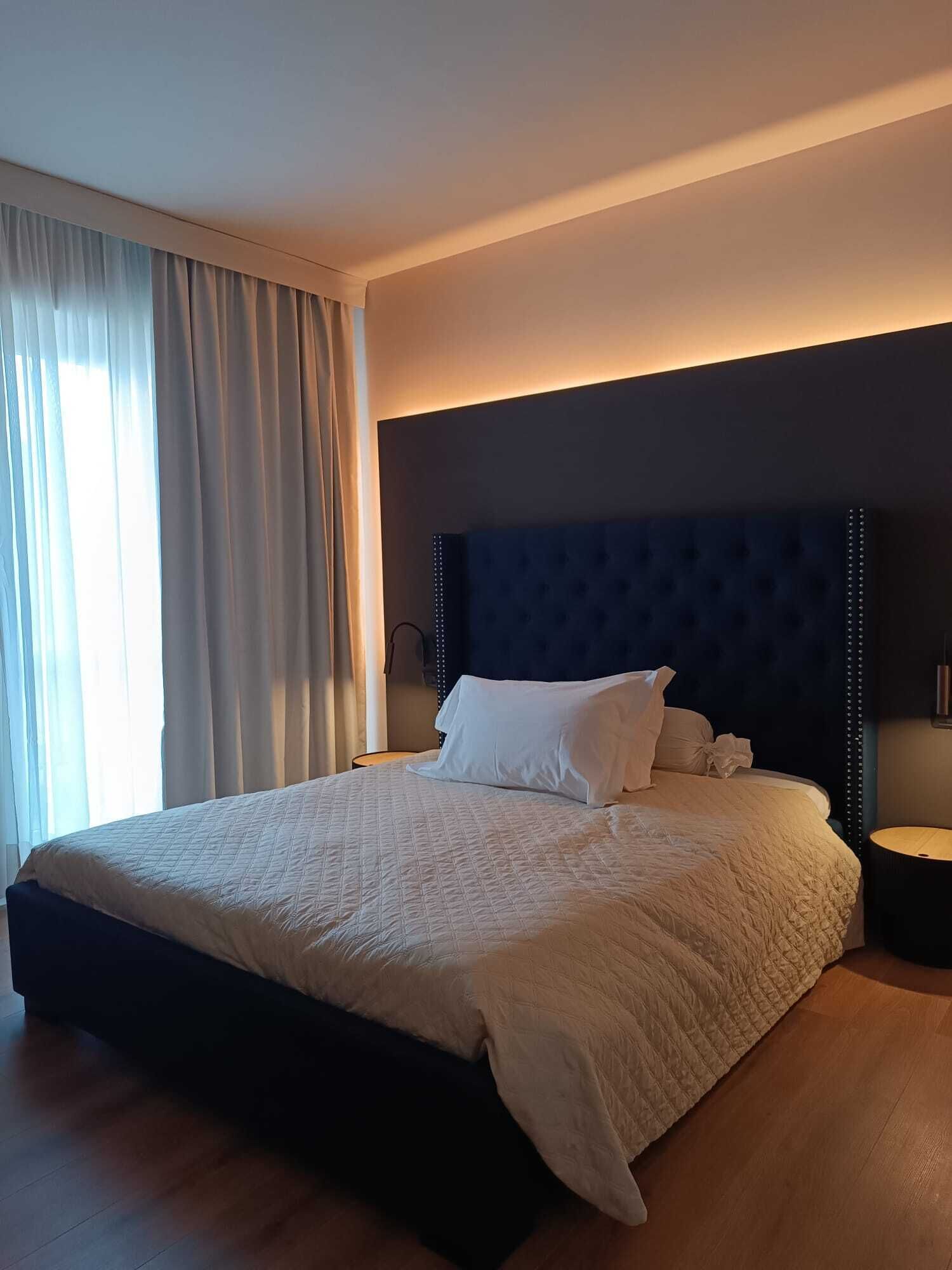 Una cama bien hecha con un cabecero oscuro y ropa de cama blanca en una habitación luminosa con un ventanal y cortinas.