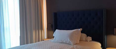 Una cama bien hecha con un cabecero oscuro y ropa de cama blanca en una habitación luminosa con un ventanal y cortinas.