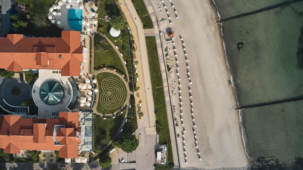 Vista aérea de una zona costera con playa, un jardín circular y un edificio de techo rojo.