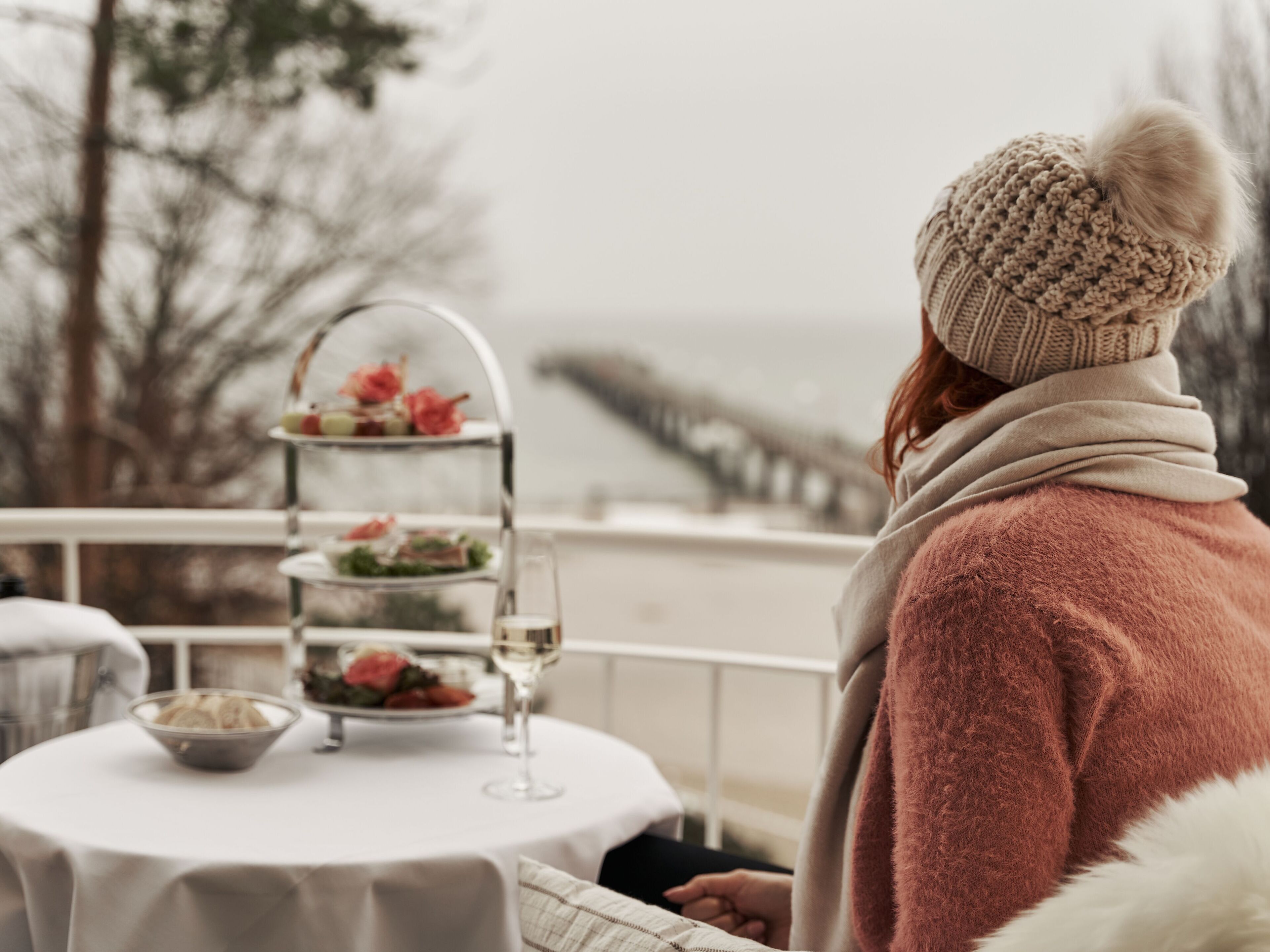 A person seated at a table with a three-tiered dessert stand, a wine glass, and a bowl of fruit. The setting appears to be an outdoor dining area with a view of a bridge in the background.