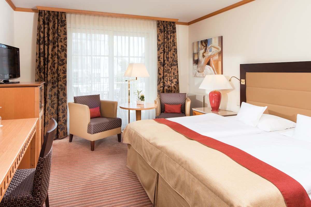 Une chambre d’hôtel comprenant un lit, deux fauteuils, un bureau avec une télévision, une lampe et un tableau au mur.