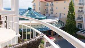Un balcón con una silla de mimbre, baranda de vidrio y vista a un edificio con varios balcones y una estructura de techo de cristal.