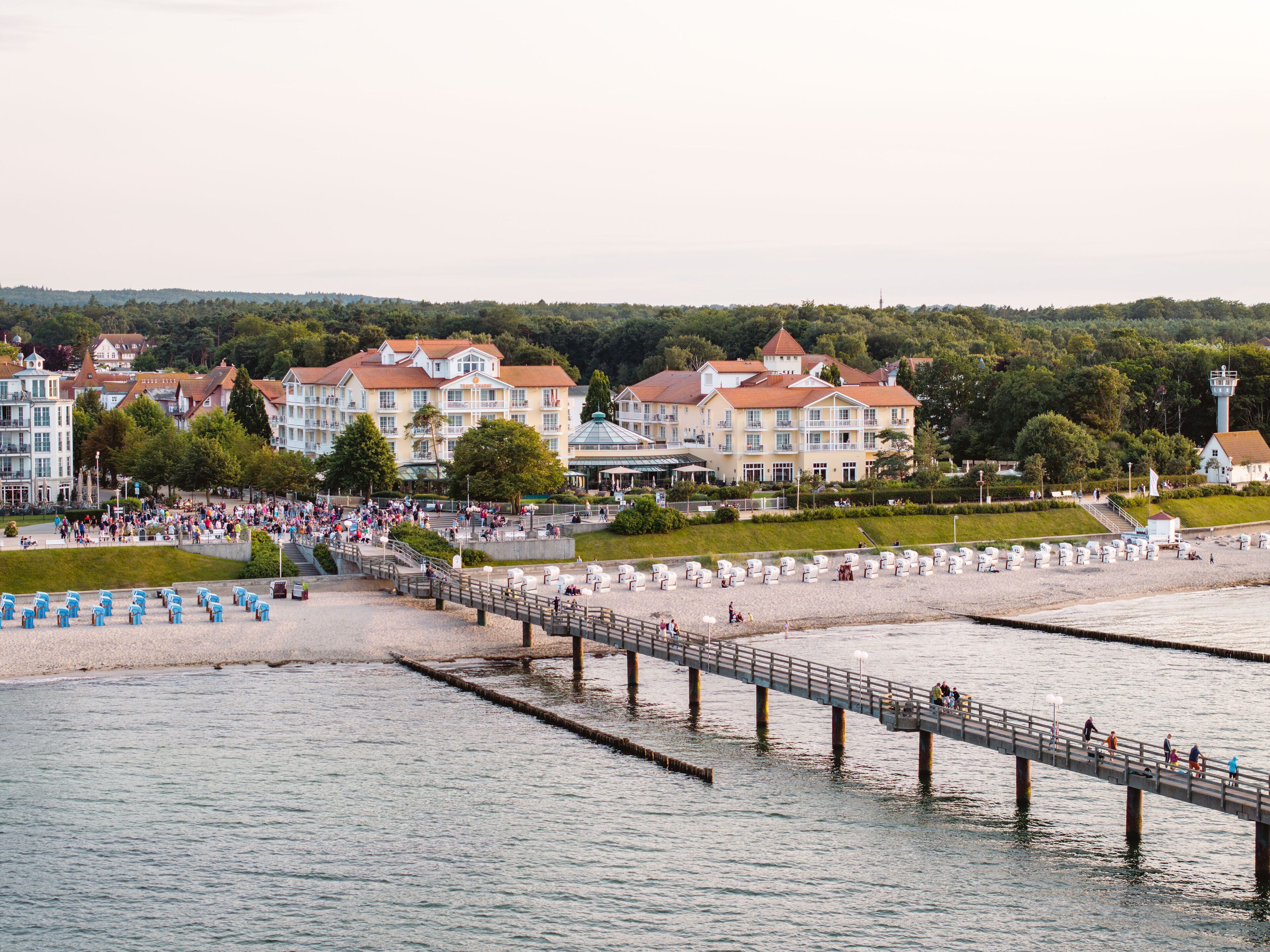 En strandpromenad längs vattnet med en brygga, en strand med solstolar och en rad byggnader med balkonger.