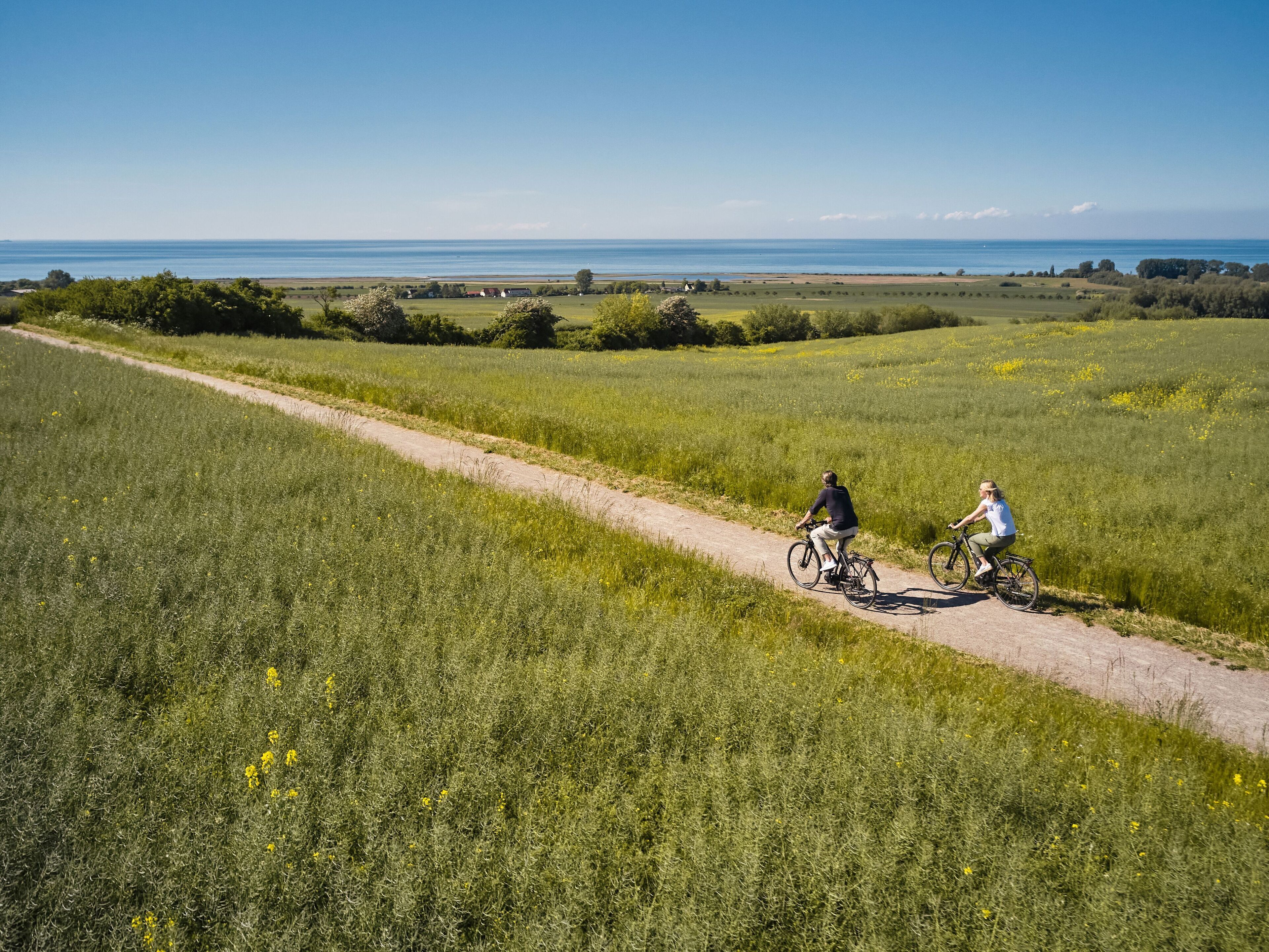 Un chemin de campagne où l’on aperçoit deux cyclistes, entourés de champs et avec une vue lointaine sur la mer.