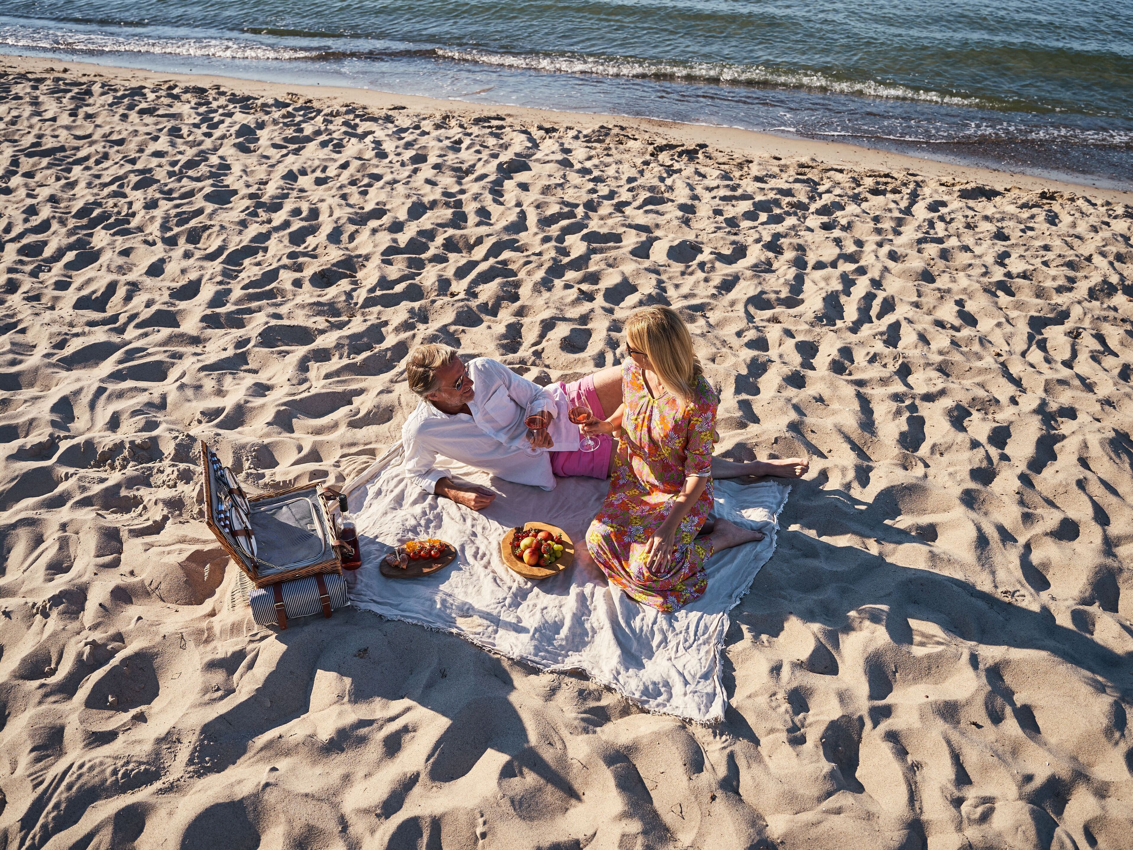 Una playa con dos personas sobre una manta, una cesta de picnic y un enfriador.