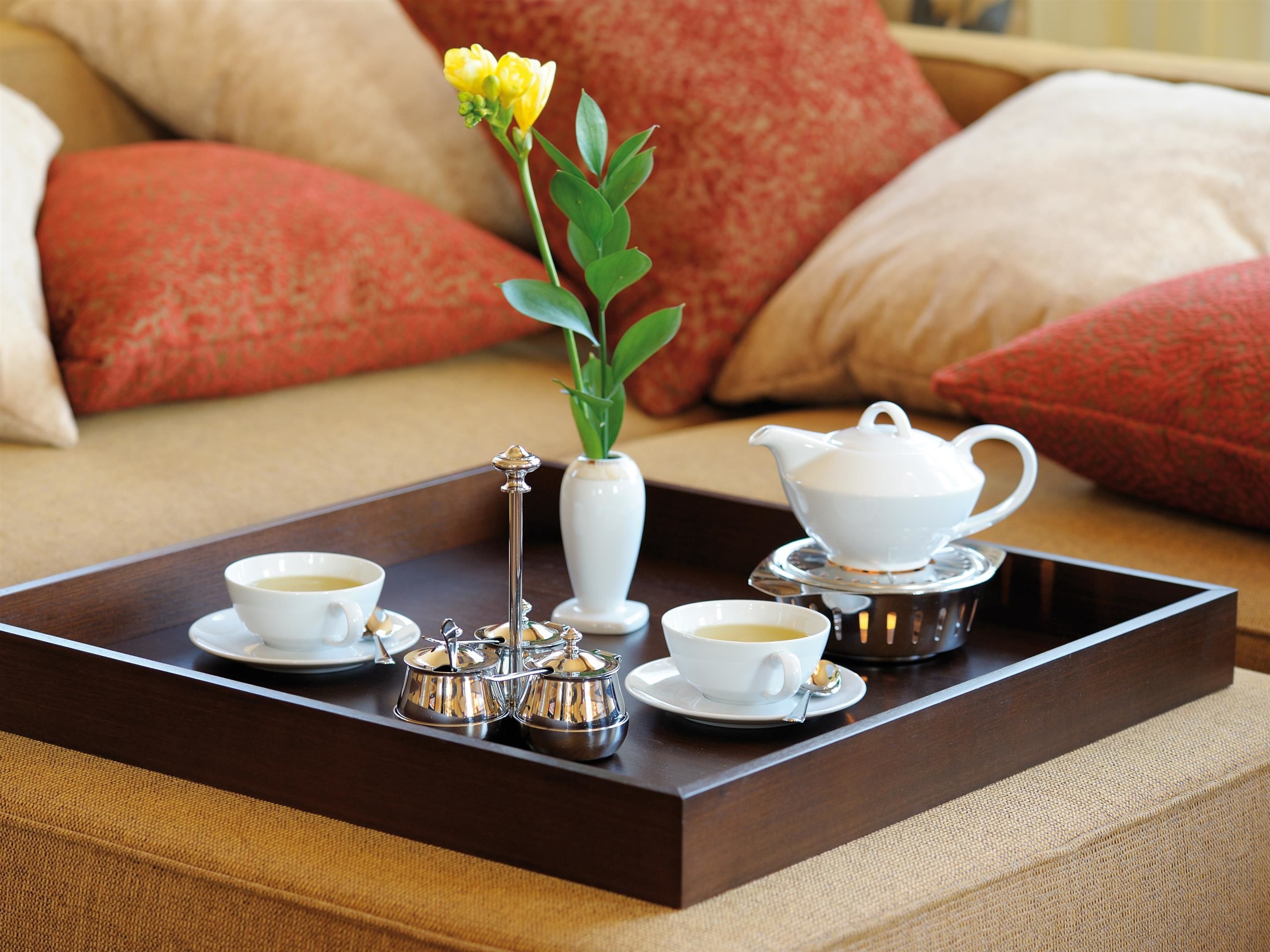 A tray with tea service on a wooden table, a white vase with a yellow flower, and a white teapot.