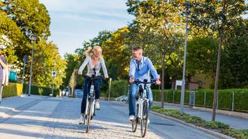 Two people riding bicycles on a paved path with trees and a fence on the side.