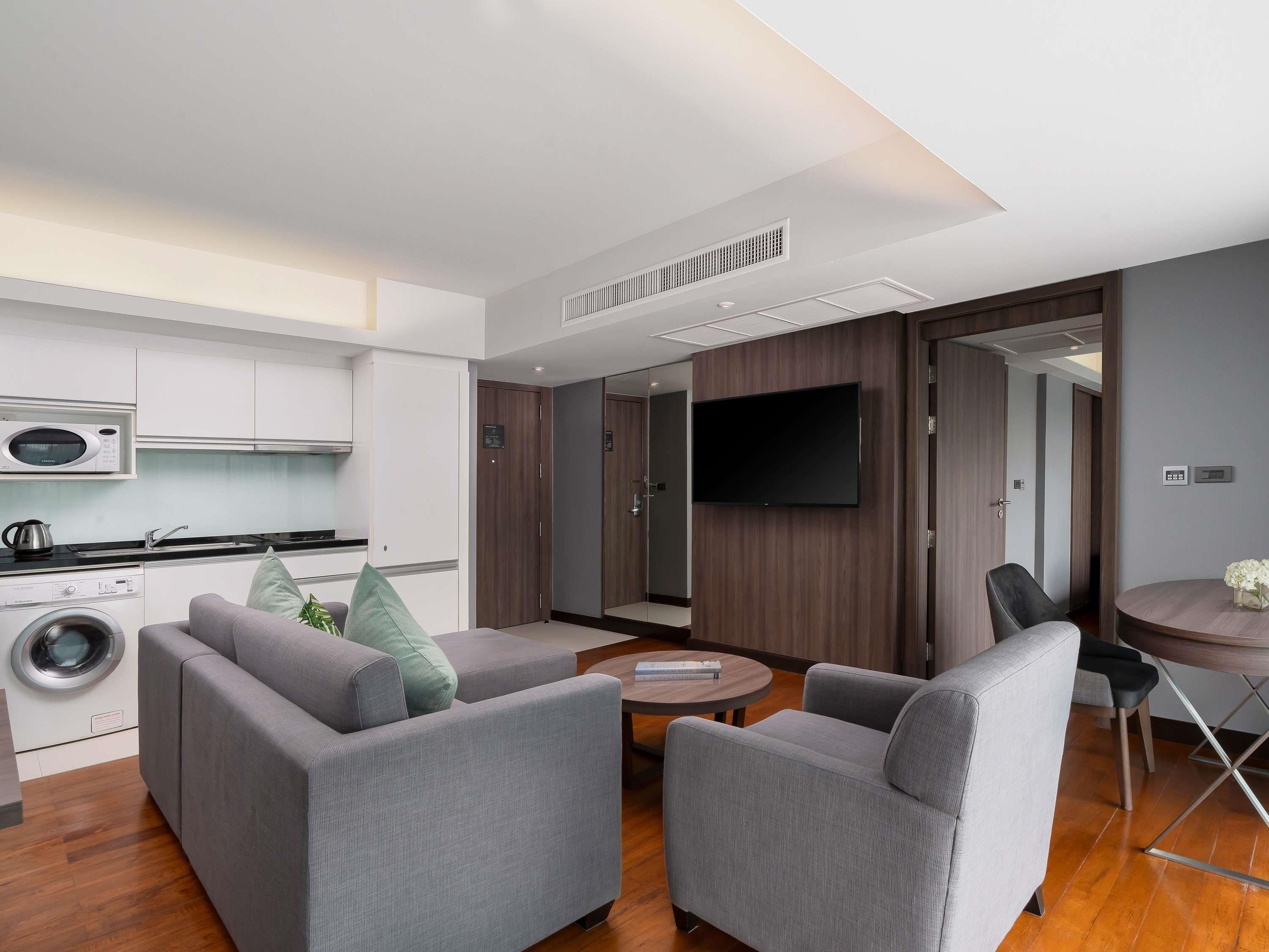 A modern living room with grey sofas, a flat-screen TV, a round coffee table, and a kitchen area with a microwave and washing machine.