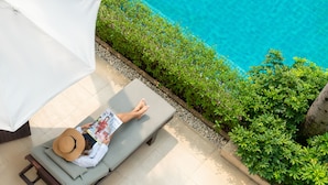 A person lying on a sunbed by a poolside, with a white umbrella and a hat.