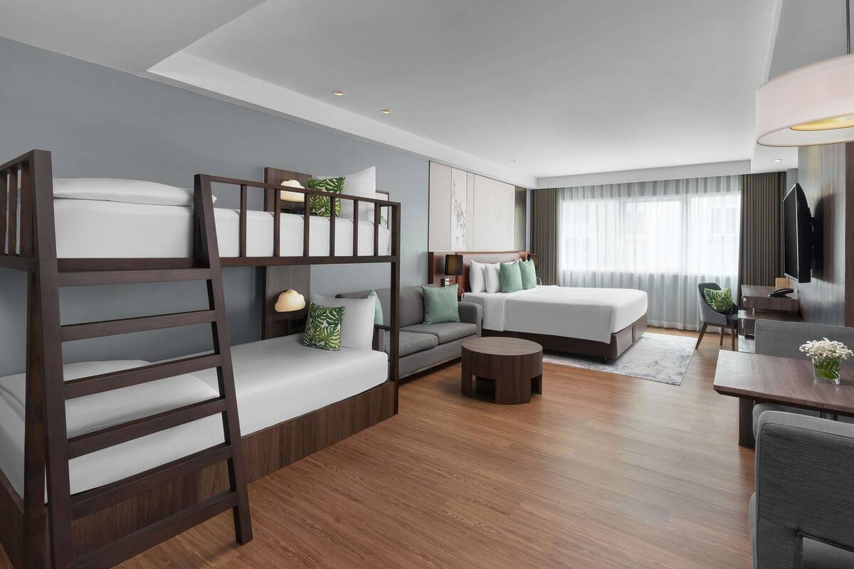 A modern hotel room with a bunk bed, a sofa, a desk, a TV, and a large window with curtains.