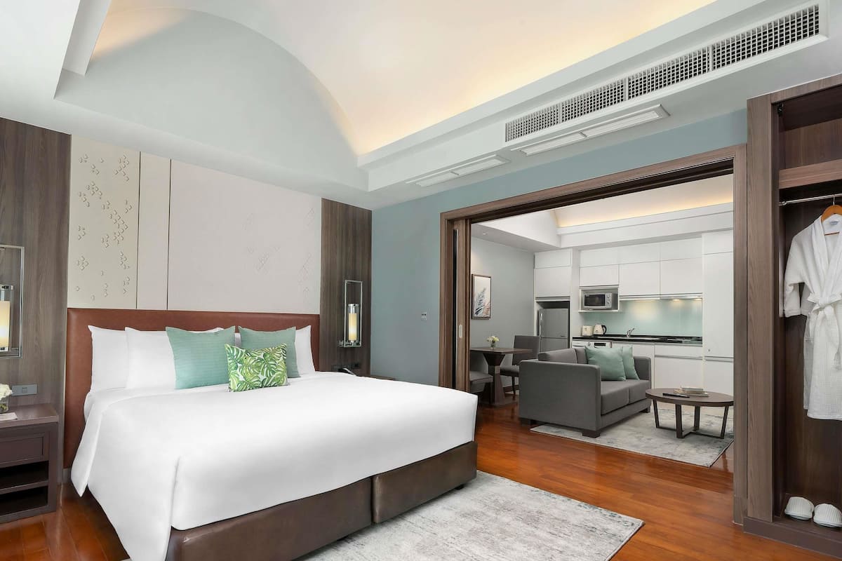 A modern hotel room with a large bed, a small kitchenette, a seating area, and a walk-in closet.
