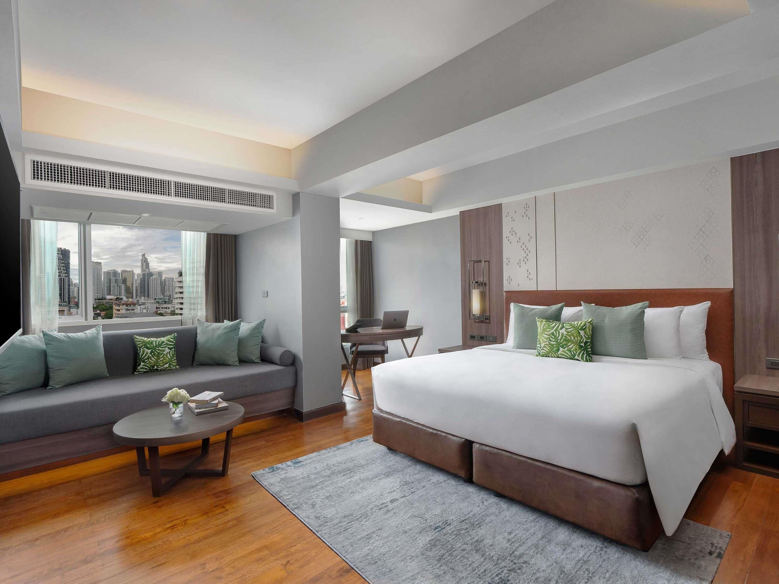 A modern hotel room with a large bed, a sofa, a dining table, and a city view.