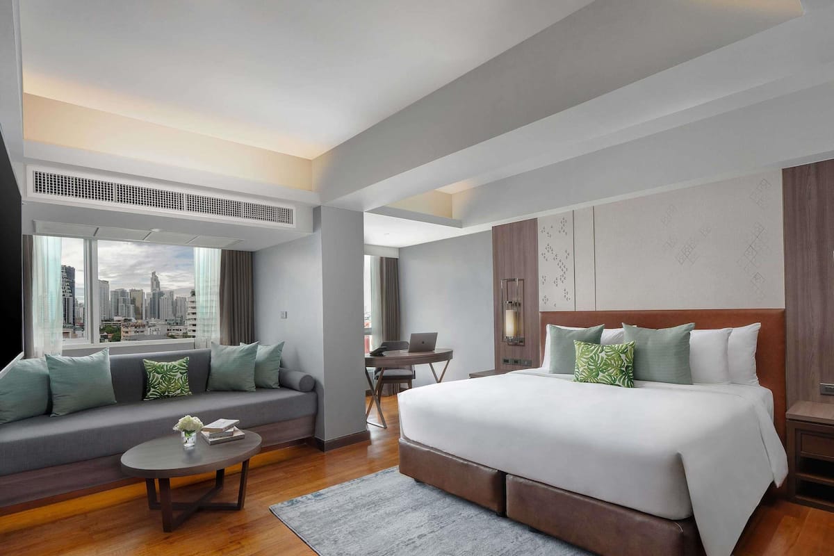A modern hotel room with a large bed, a sofa, a dining table, and a city view.