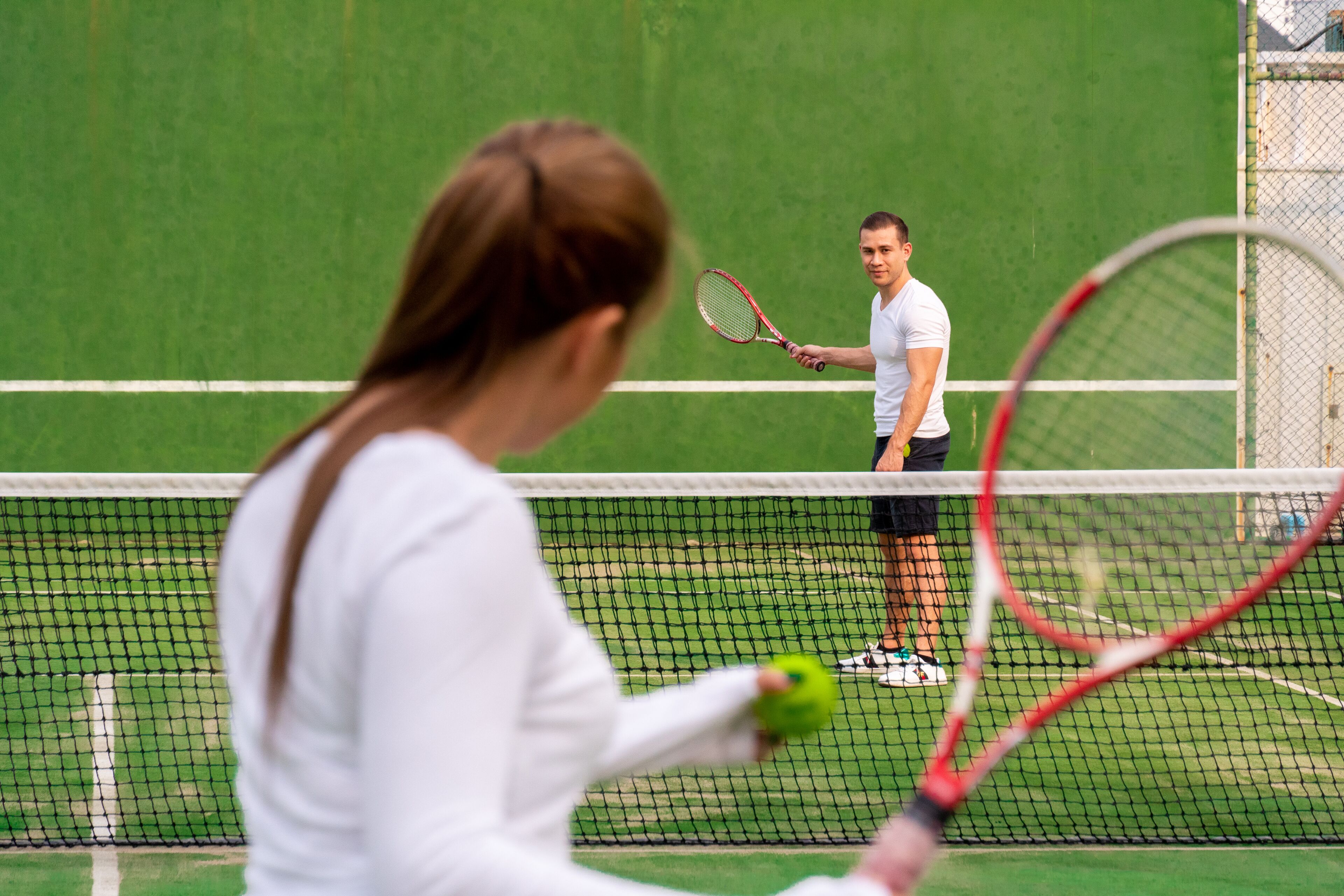 A tennis court with a net in the middle, a player in a white shirt, and another player in the background.