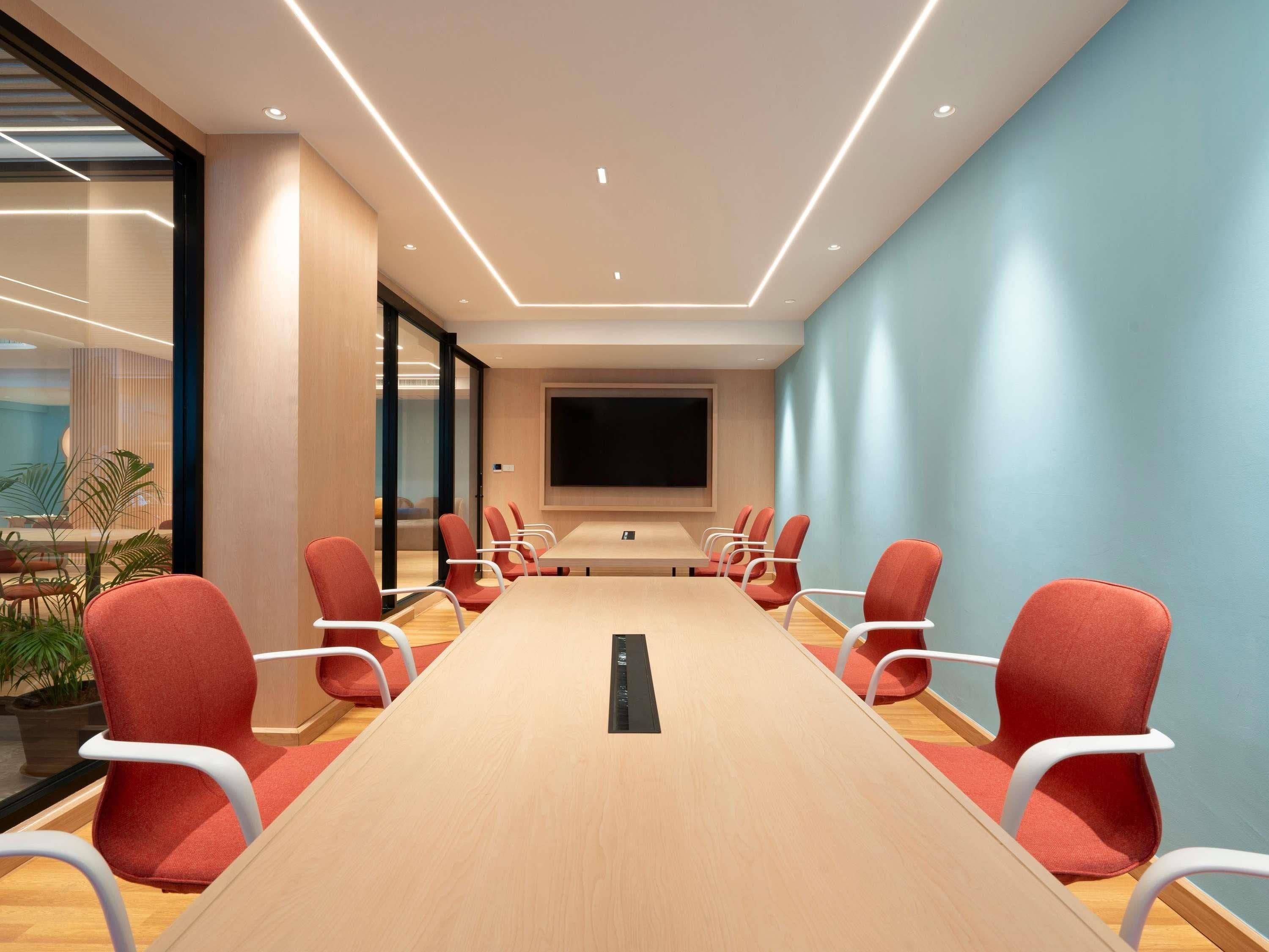 A conference room with a long table, red chairs, a large screen, and a glass wall.
