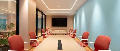 A conference room with a long table, red chairs, a large screen, and a glass wall.