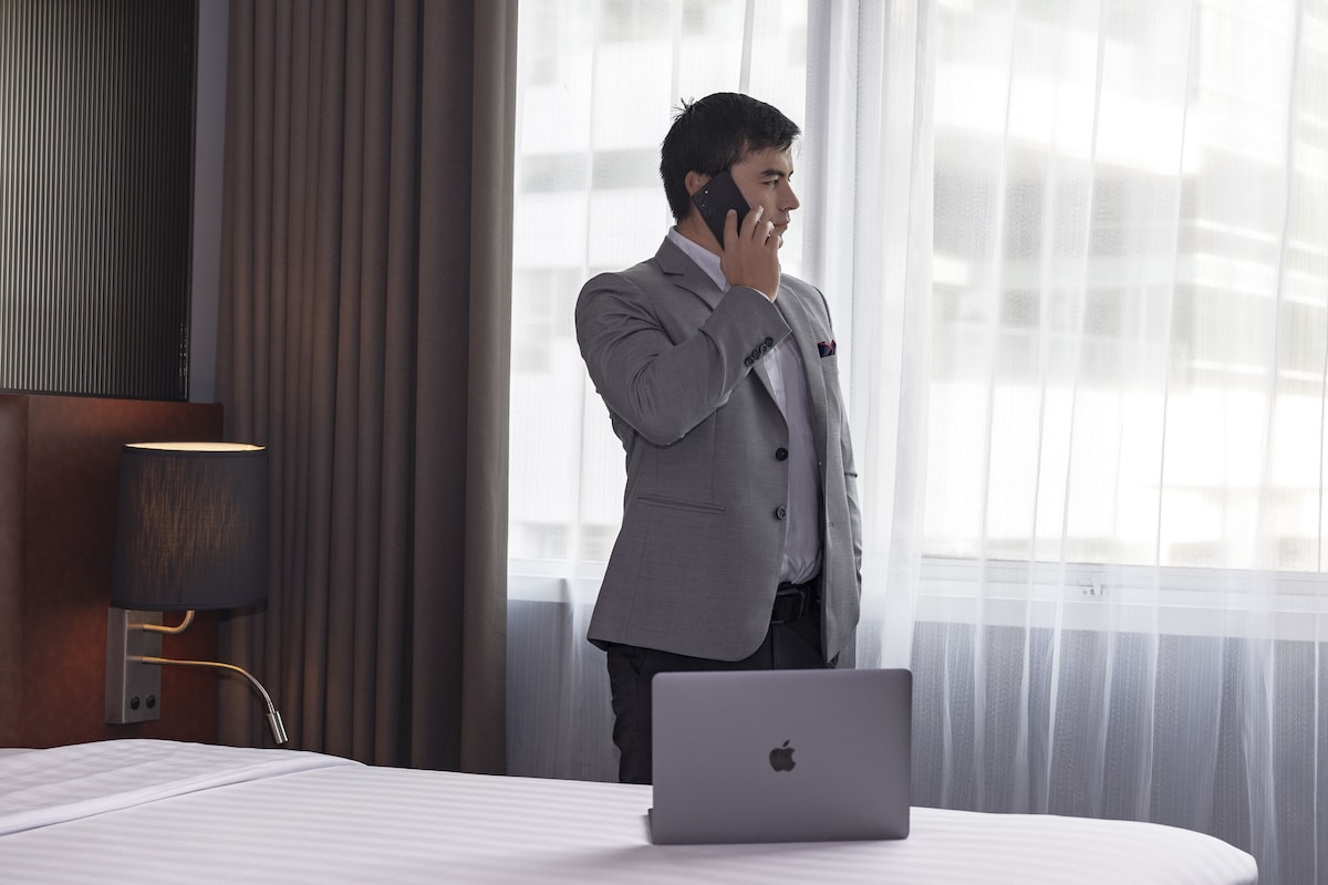 A hotel room with a bed, a laptop, and a person talking on a phone.