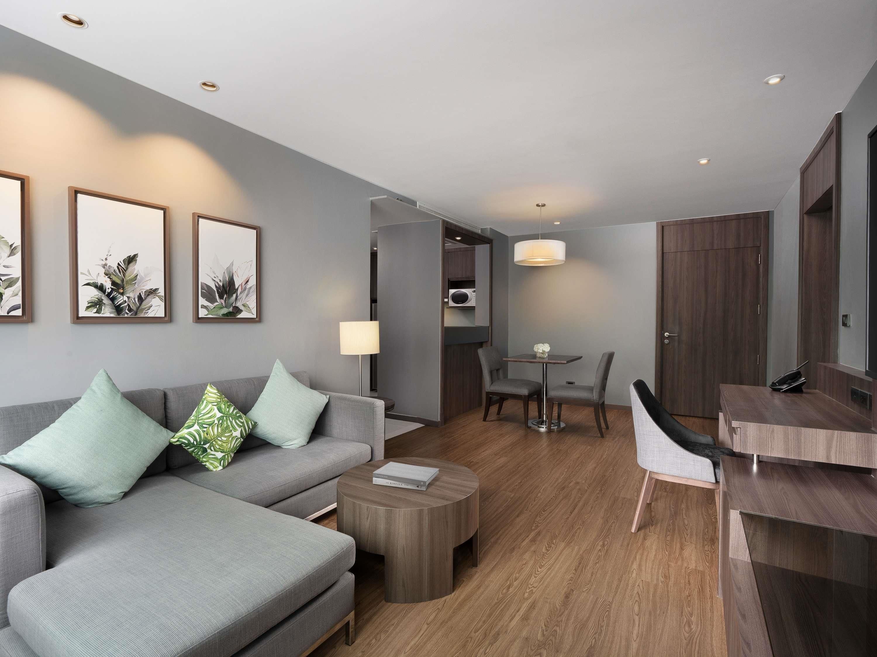 Una habitación de hotel moderna con un sofá gris, una pequeña mesa redonda, un área de comedor con una mesa y sillas, y un armario.