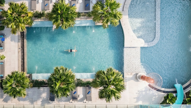 An aerial view of a swimming pool with a slide, surrounded by palm trees and lounge chairs. - Novotel Living Bangkok Sukhumvit Legacy (Bangkok)