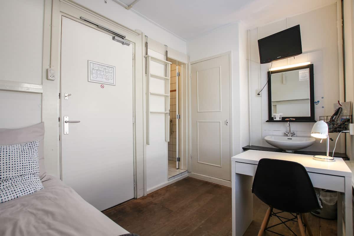 Una habitación de hotel con una cama, un escritorio con una silla, una lámpara de escritorio, un televisor sobre el escritorio y un baño con un lavamanos y un espejo.