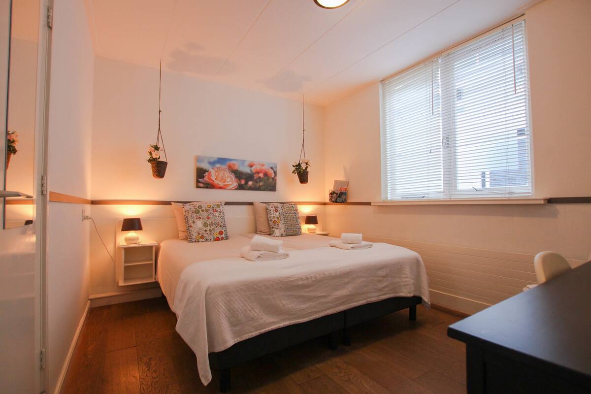 Un dormitorio ordenado con una cama grande, mesitas de noche con lámparas, una ventana con persianas y un cuadro colgado en la pared.