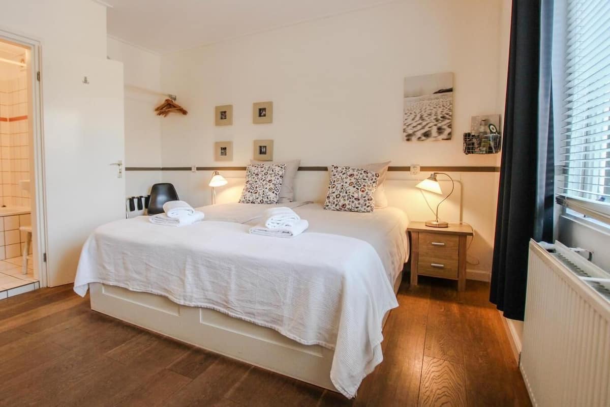 Un dormitorio con una cama grande, mesitas de noche, un radiador y una ventana con persianas.
