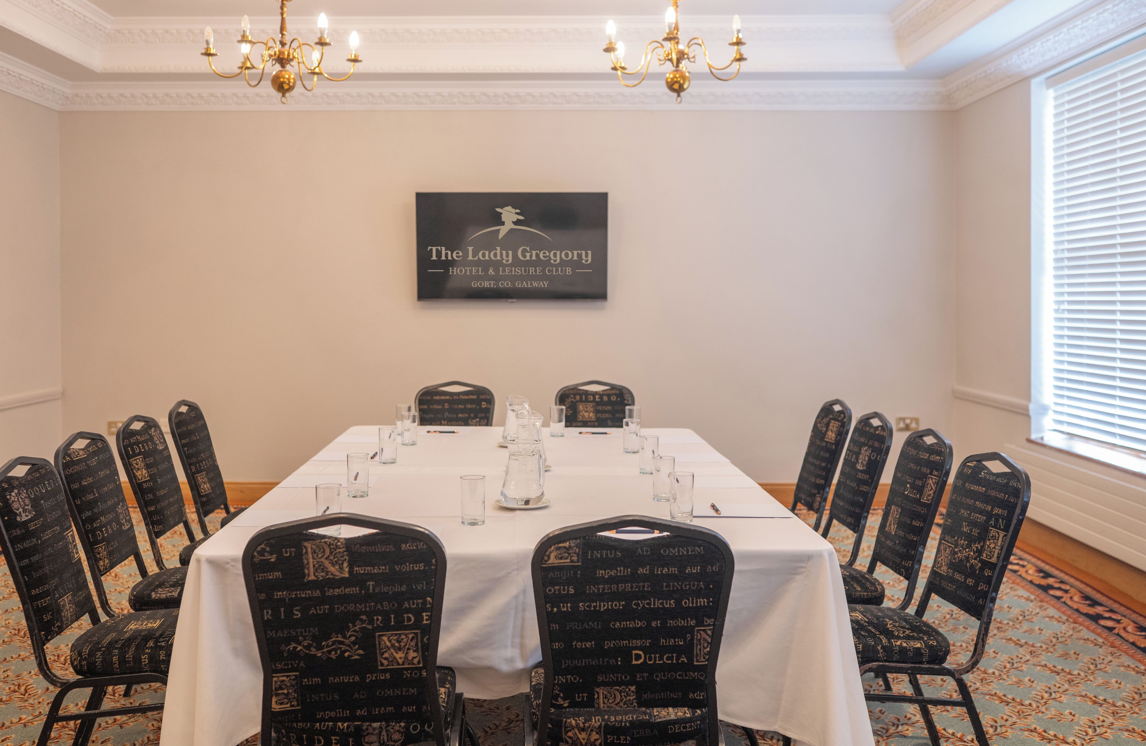 A conference room with a long table, chairs with decorative backrests, and a chandelier. A sign on the wall reads 'The Lady Gregory Hotel & Leisure Club.'