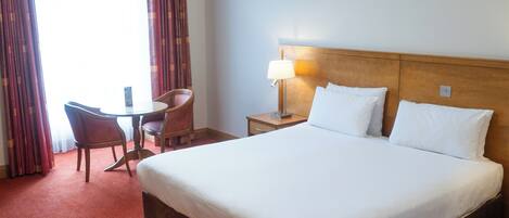 A hotel room with a large bed, a bedside table with a lamp, a chair, and a small table.