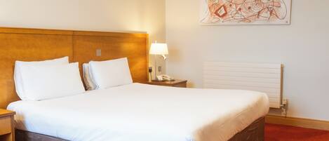 A hotel room with a large bed, a nightstand with a lamp, a wall-mounted artwork, and a radiator.