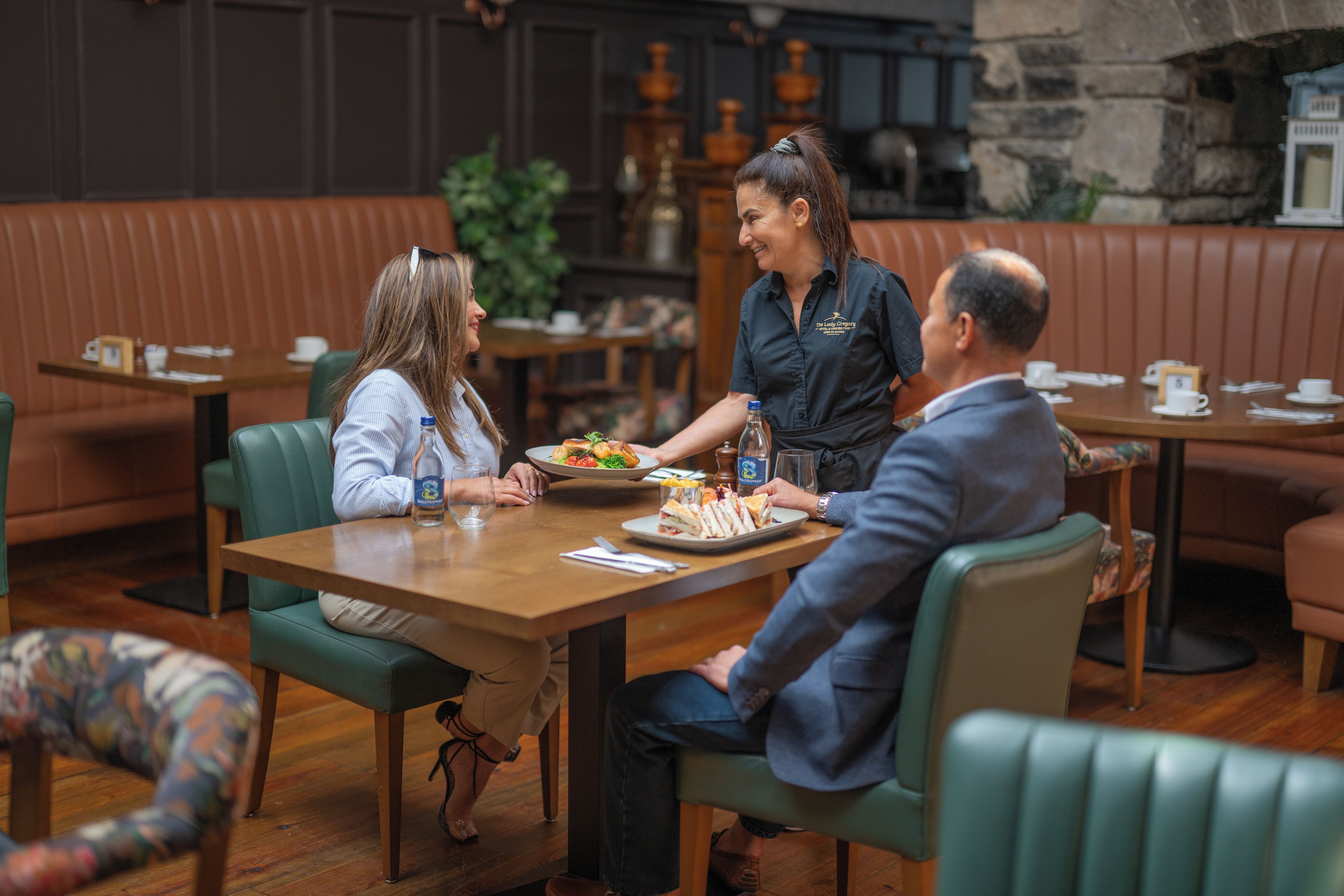 Une salle de restaurant avec une table en bois, des chaises à revêtement vert et une serveuse qui apporte les plats.
