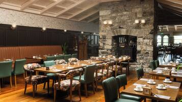 Un restaurante con mesas de madera, sillas tapizadas de verde, una chimenea de piedra y un techo alto con molduras decorativas.