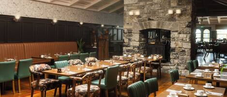 Un restaurante con mesas de madera, sillas tapizadas de verde, una chimenea de piedra y un techo alto con molduras decorativas.