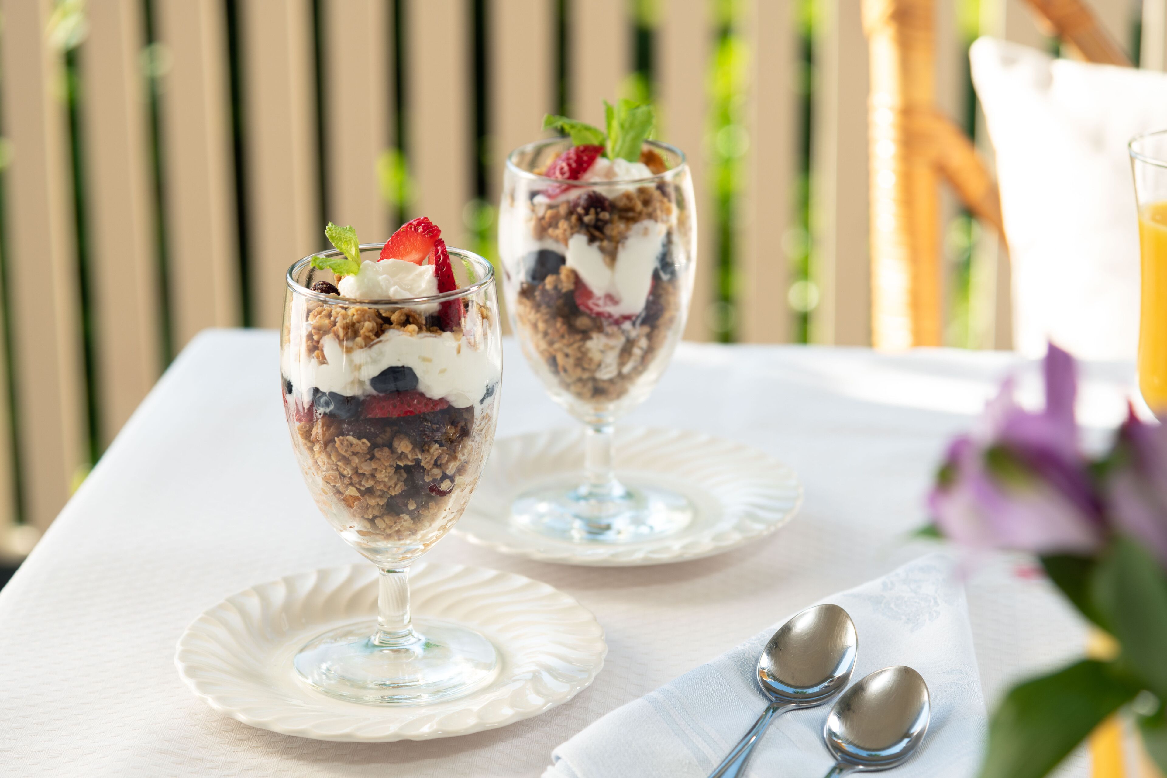 Two dessert glasses on white plates with a white tablecloth, accompanied by two spoons and a napkin.