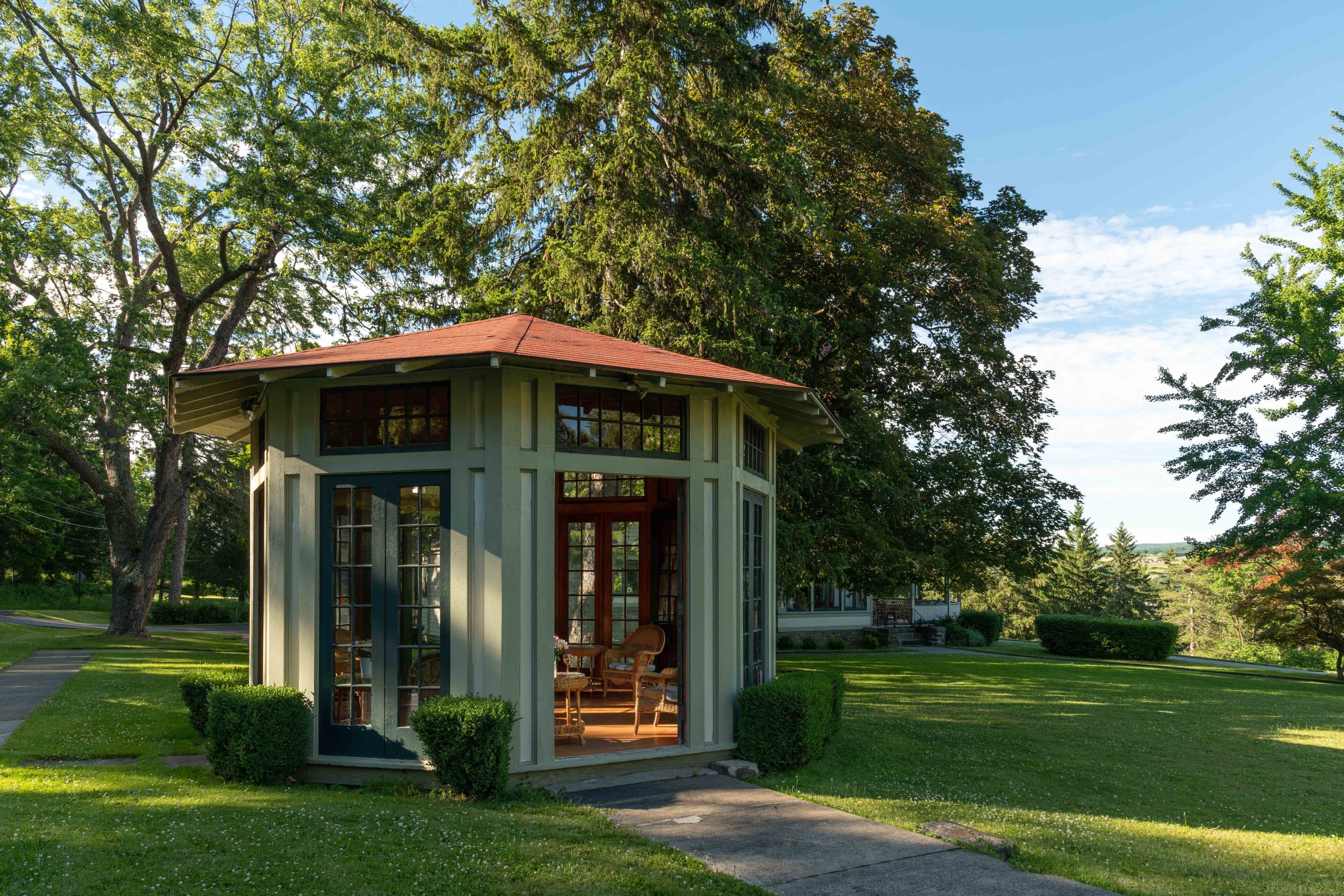 A small, single-story structure with a red roof, large windows, and a door, surrounded by a well-maintained lawn and trees.