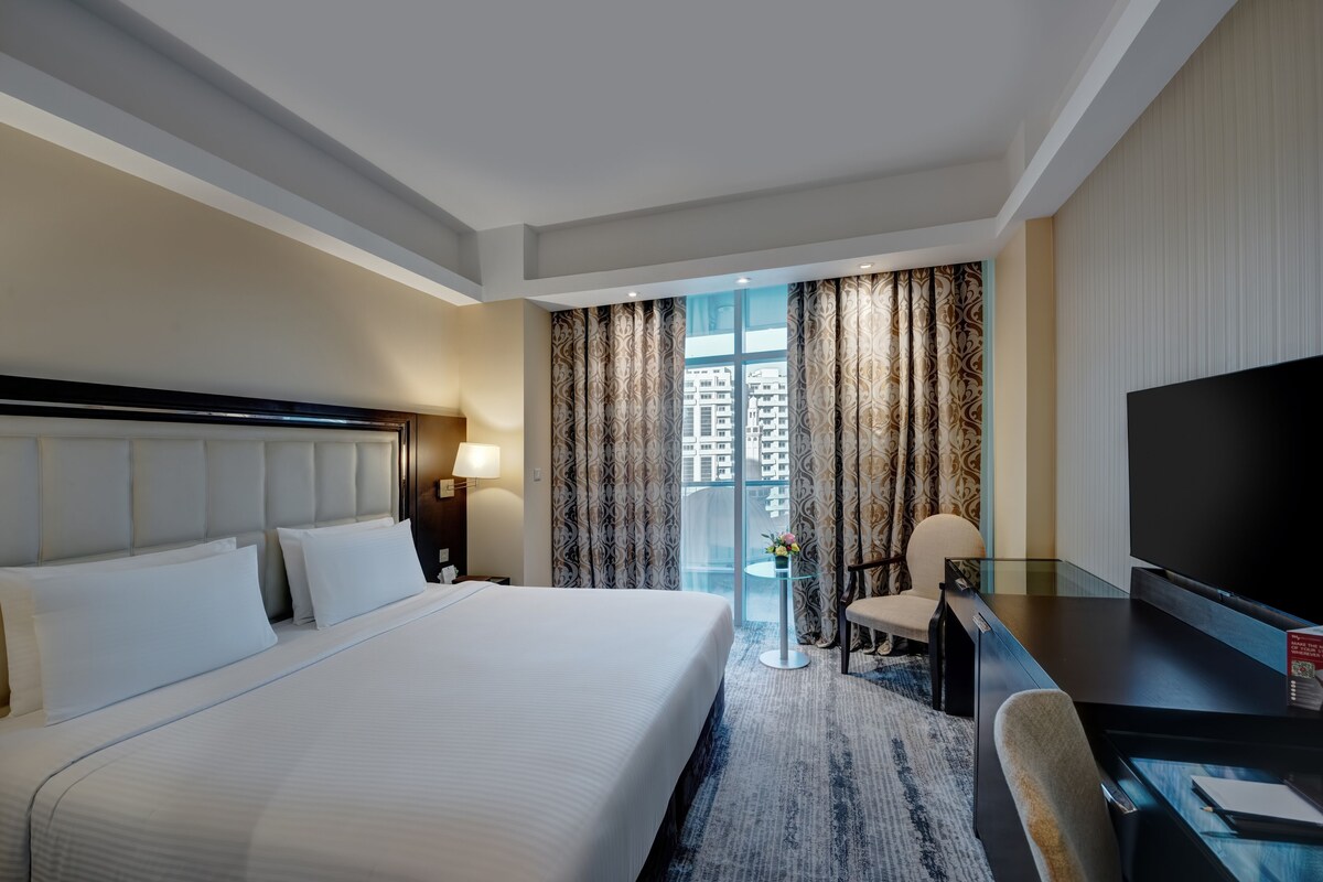 A hotel room with a large bed, a desk with a TV, a chair, and a view of buildings through the window.