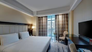 Habitación de hotel con una cama grande, un escritorio con televisor, una silla y vistas a edificios a través de la ventana.