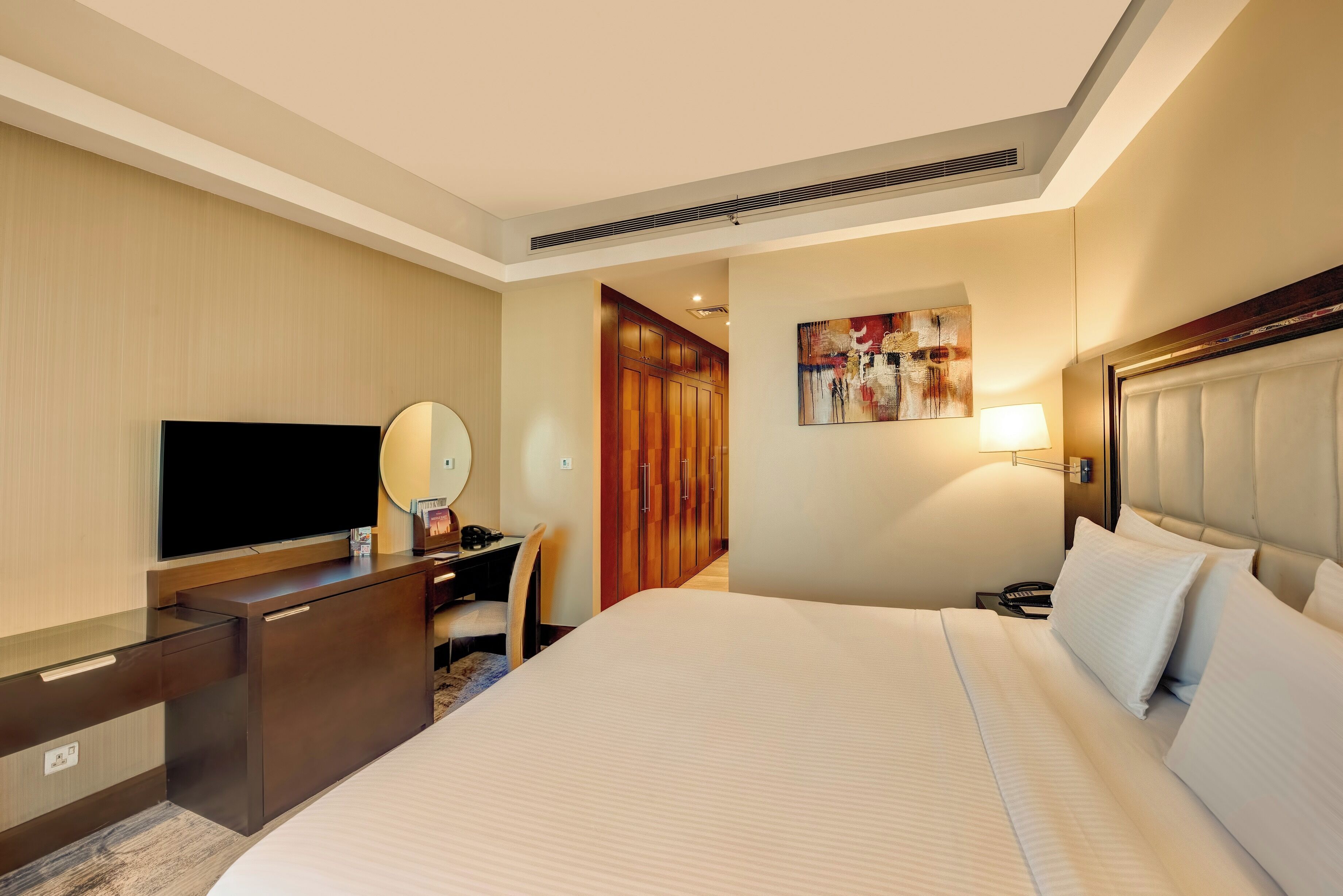 Une chambre d’hôtel avec un grand lit, un bureau avec une chaise, une télévision, un miroir, un tableau et une table de chevet avec une lampe.