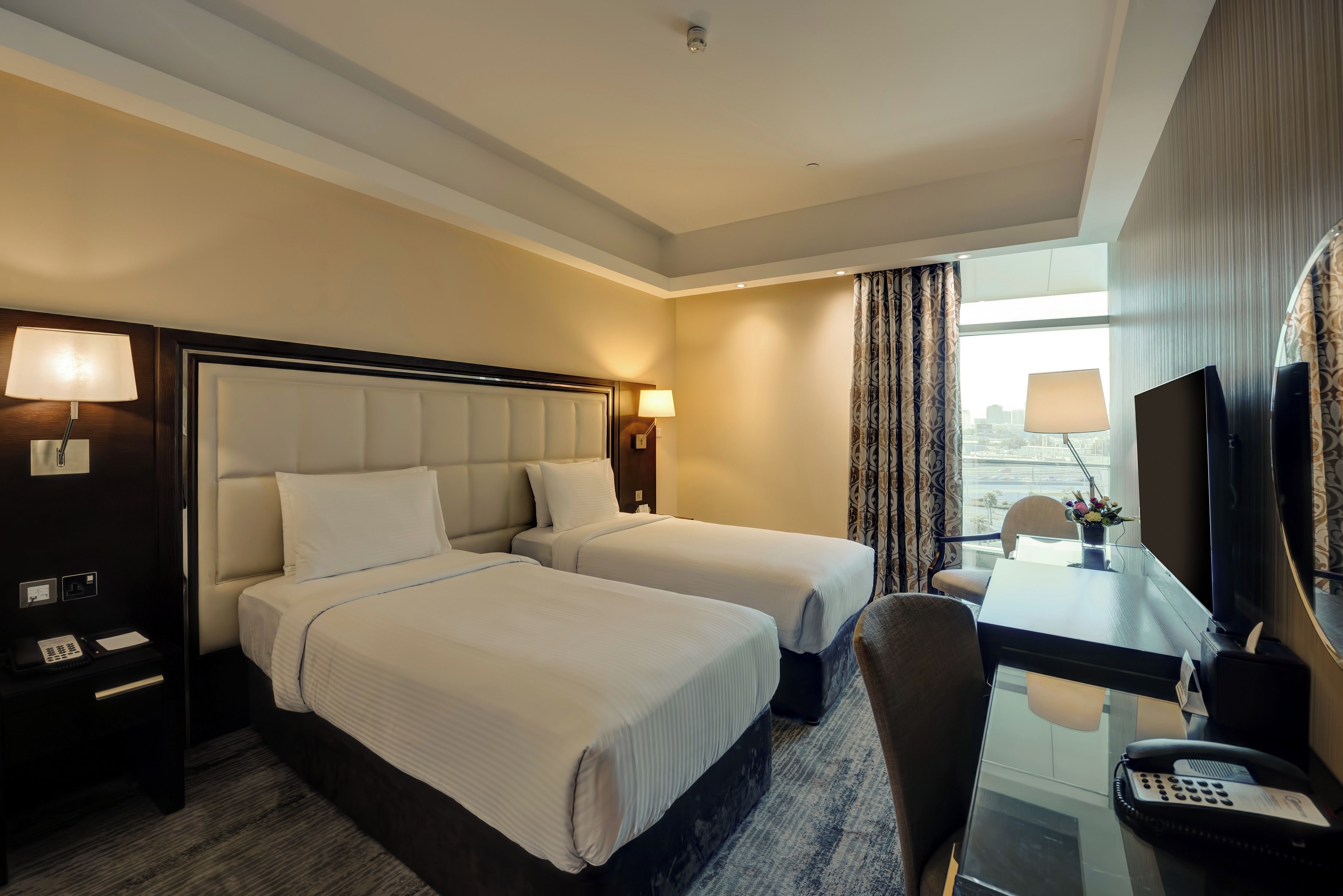 A hotel room with two beds, a desk with a telephone, and a large window offering a city view.