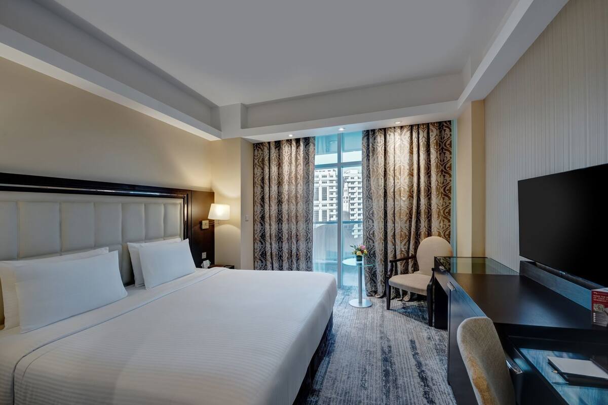 A hotel room with a large bed, a desk with a chair, a TV, and a view of buildings through the window.