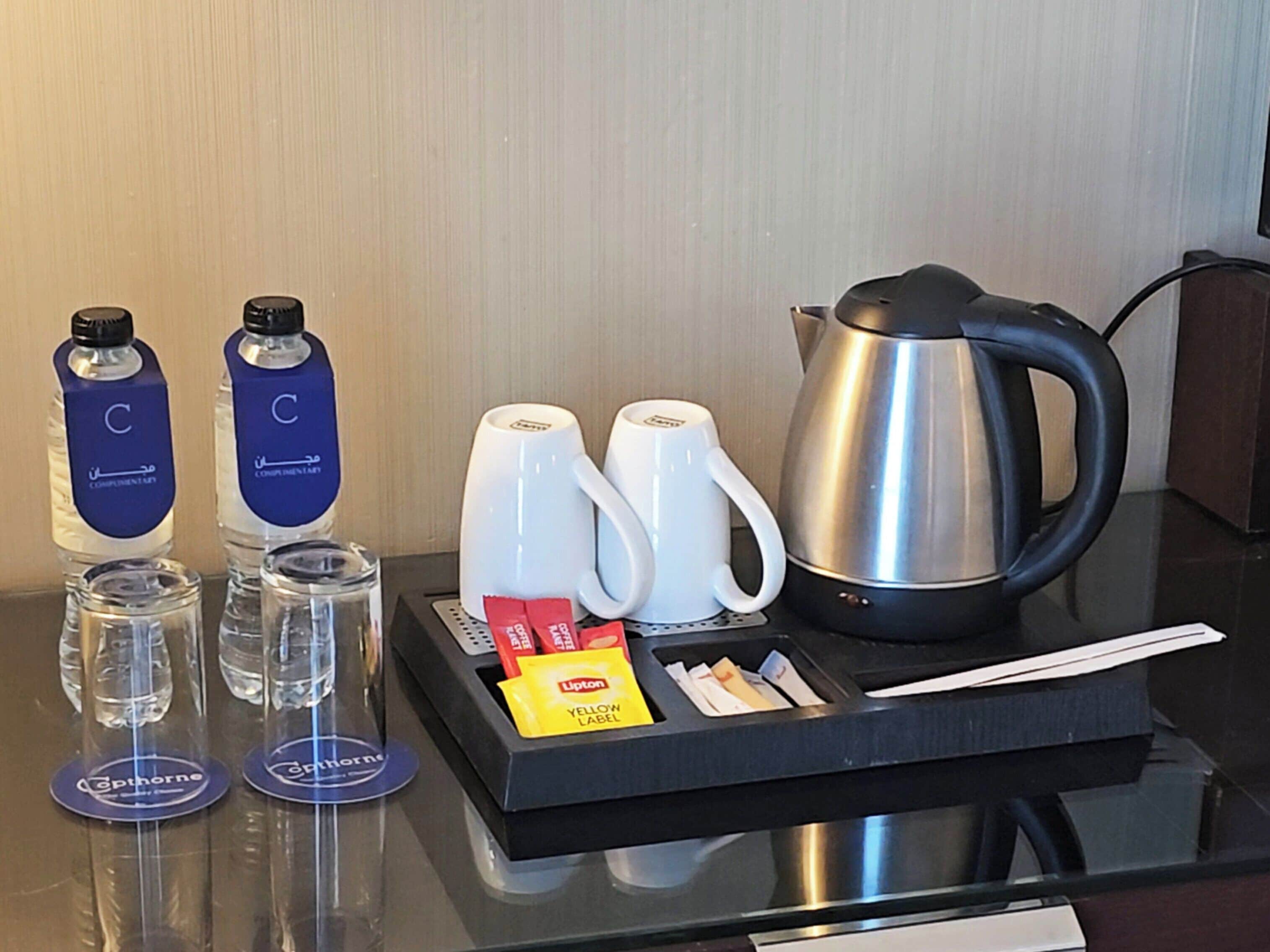 A hotel room's minibar with bottled water, coffee mugs, a kettle, and a tray with tea bags and a yellow label.